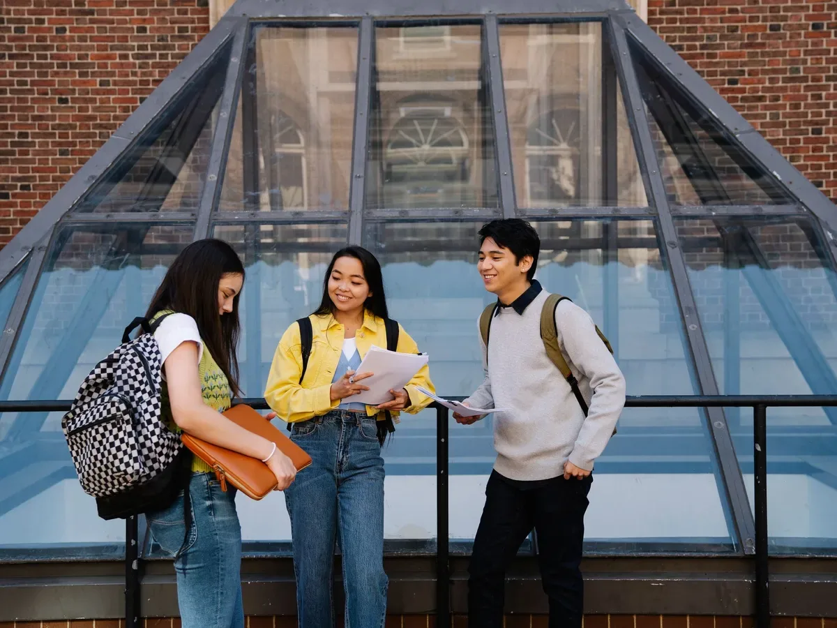 ثلاثة طلاب يتحدثون معاً حول أوراق مشتركة أمام مبنى جامعي